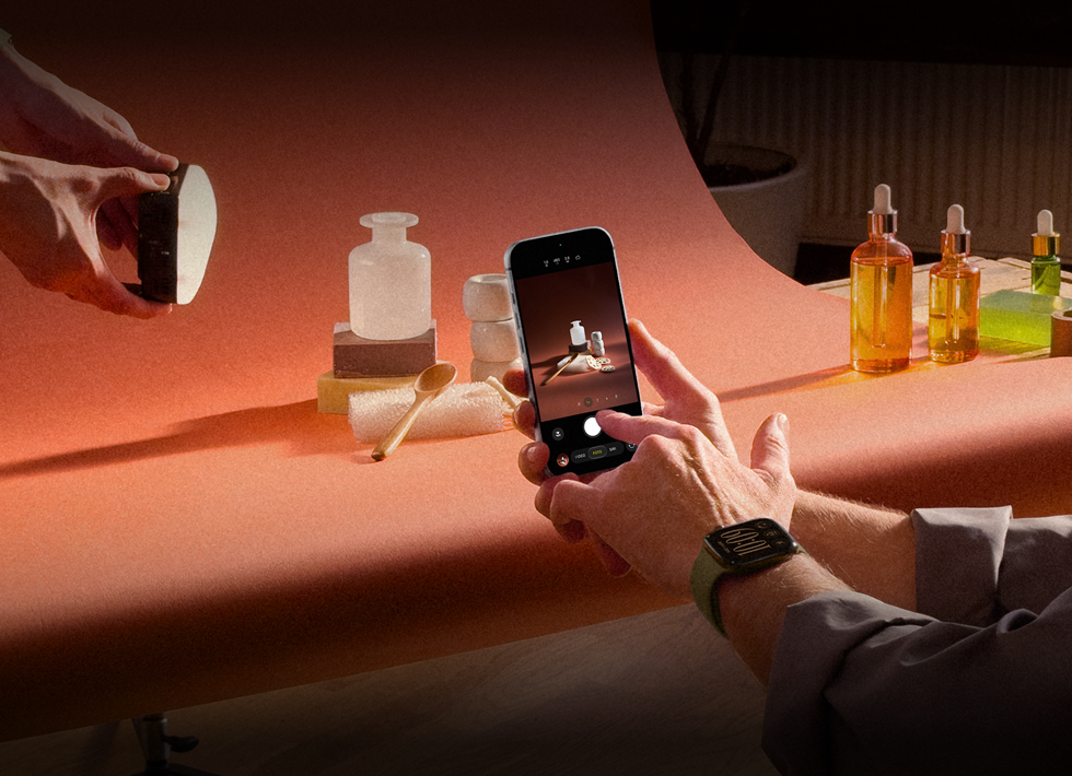 Una sesión de fotos en un lugar de trabajo, una persona usa un iPhone para fotografiar productos sobre un fondo, otra persona sostiene un foco